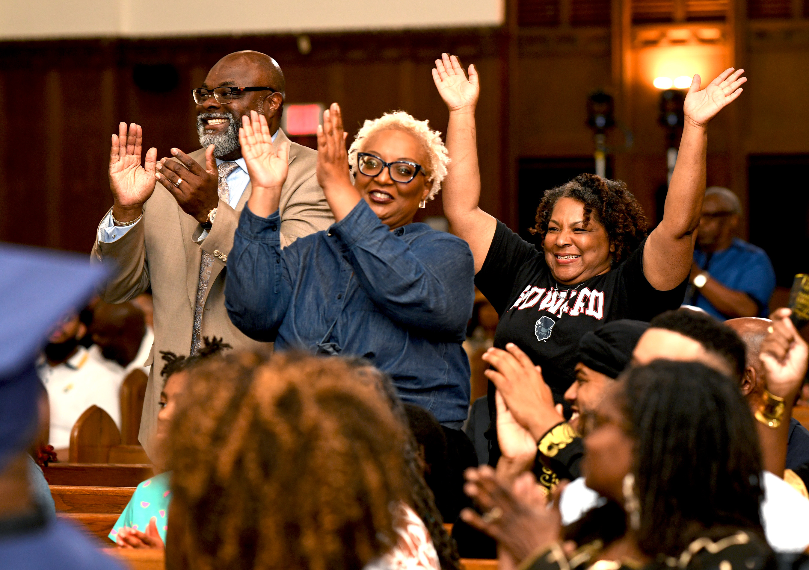 Three individuals stand in the benches of a crowded church: a man in a tan suit with matching colorful tie, a woman in blue denim, and a woman in a black Howard tshirt with a bison mascot, stand smiling and cheering with arms raised among others smiling and clapping in the crowd.