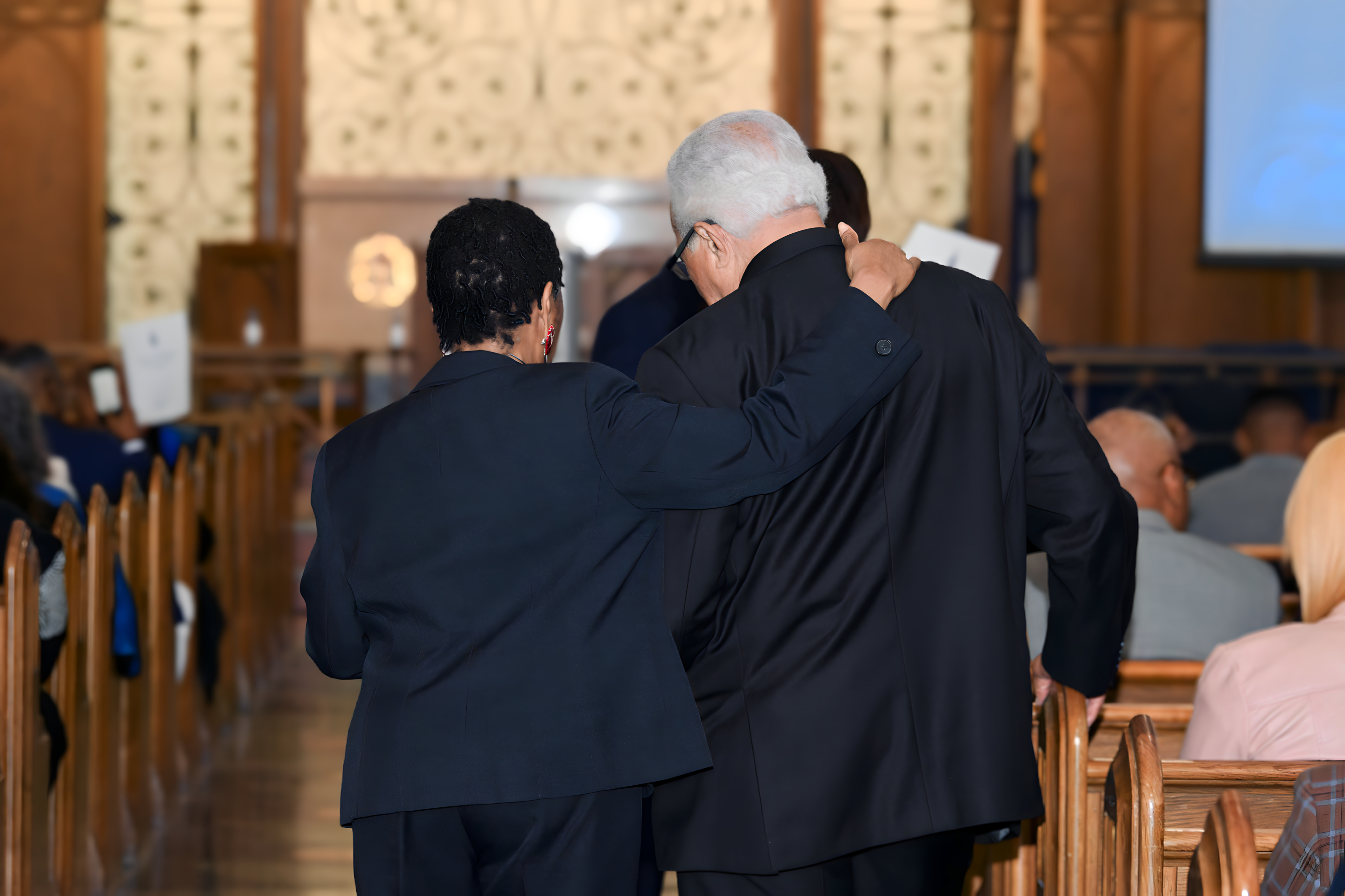 Inside of a church, two people in dark suits embrace in fellowship as they walk up the hall. Ornate wooden benches with seated guests are on either side of them. The edge of a screen can be seen up front.