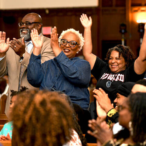 Three individuals stand in the benches of a crowded church: a man in a tan suit with matching colorful tie, a woman in blue denim, and a woman in a black Howard tshirt with a bison mascot, stand smiling and cheering with arms raised among others smiling and clapping in the crowd.